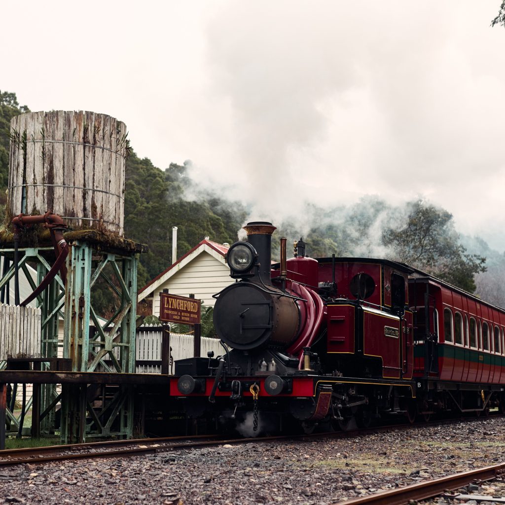 A steam locomotive waiting by the station's platform, a sign displaying that the train is stopped at Lynchford station. Nearby a water tank in the foreground, and steam bellowing from the loco's chimney.