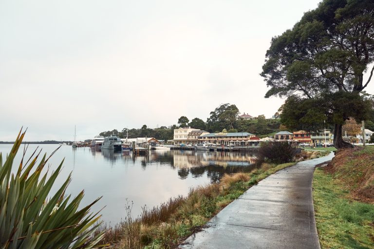 An image of the Strahan waterfront with a walking track to the right and the Macquarie Harbour to the left. Image also shows the main township of Strahan in the background along with boats that are docked.